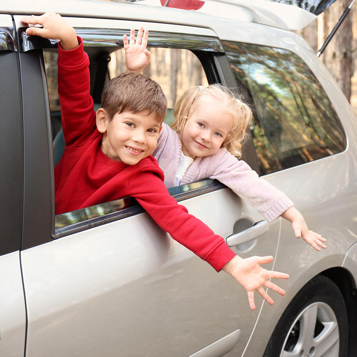 Eine Auto-Reise mit Kindern steht bevor. Ein kleiner Junge mit rotem Pullover und ein kleines blondes Mädchen sitzen auf den Rücksitzen eines parkenden Autos und schauen aus dem geöffneten Fenster. Sie freuen sich, dass sie gleich mit dem Auto in den Urlaub fahren.