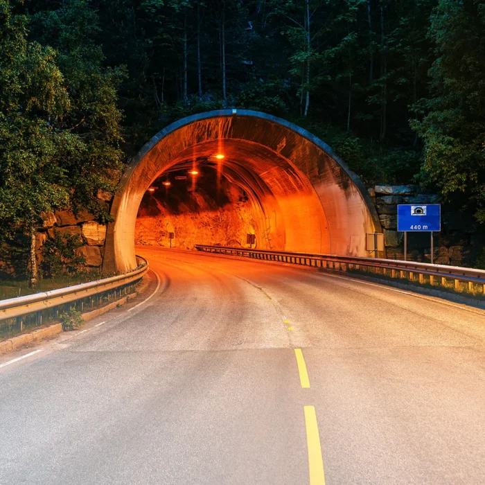 Straße führt in einen rot beleuchteten Tunnel. Der Tunneleingang ist umgeben von vielen Bäumen. Auf der rechten Seite des Tunneleingangs weist ein blaues Schild mit weißer Schrift auf die Tunnellänge von 440 Metern hin.