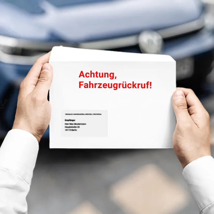 Zwei Hände halten einen Brief mit roter Aufschrift "Achtung, Fahrzeugrückruf!" in roter Schrift. Auto verschwommen im Hintergrund