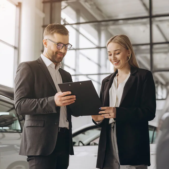 Ein Mann und eine Frau sprechen in einem Autohaus über Anforderungen von Kunden. Er zeigt ihr schwarze Klemmmappe. Sie lächelt. Drum herum stehen Fahrzeuge.