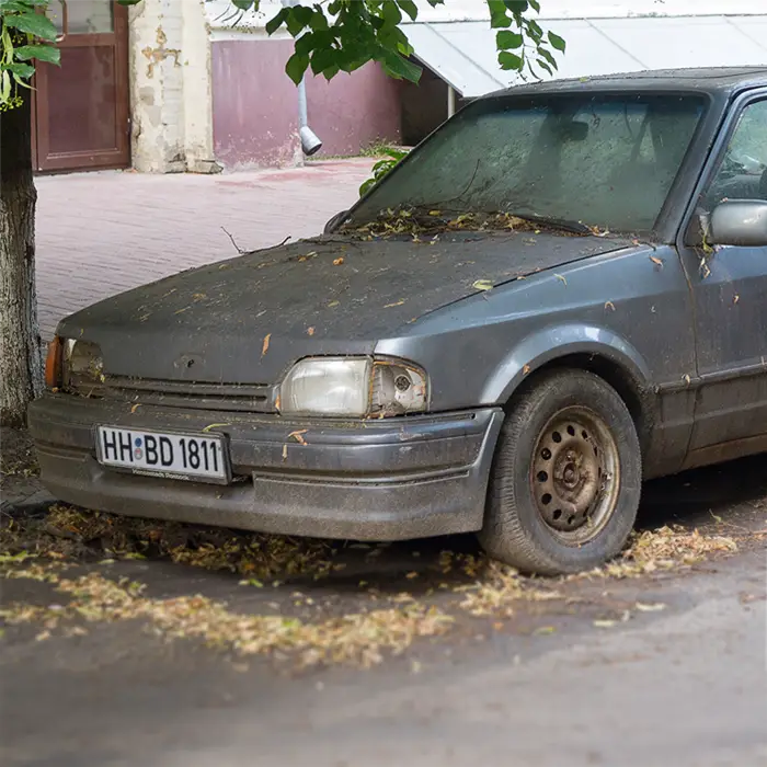 Graues Auto am Straßenrand mit sichtbaren Spuren durch Langzeitparken: Blätter/Laub und verklebte Frontscheibe, verschmutzte Karosserie, Rost an der Felge.