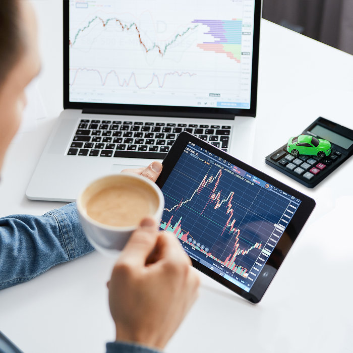 Mensch am Tisch mit Laptop und Tablet schaut sich Kurse von Auto-Aktien an und trinkt eine Tasse Kaffee