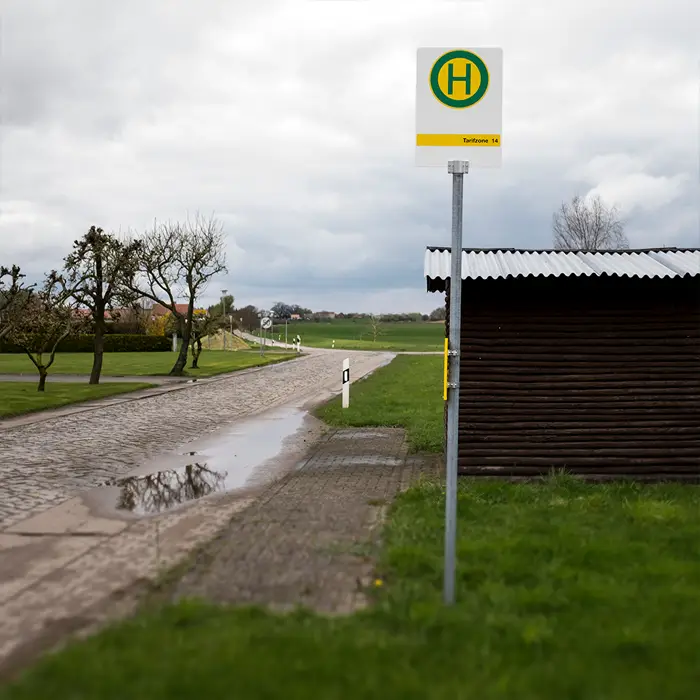 Bushaltestelle auf dem Land. Haltestellenschild auf einem Mast. Wartehäuschen aus Holz mit Dach dahinter.Auf der Straße aus Kopfsteinpflaster ist eine große Pfütze. Wiese, Felder und einzelne Bäume.