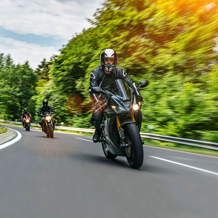 Ein Mann auf einem schwarzen Motorrad fährt auf einer asphaltierten Straße. Zwei weitere Motorräder folgen ihm. m Straßenrand stehen viele grüne Bäume.