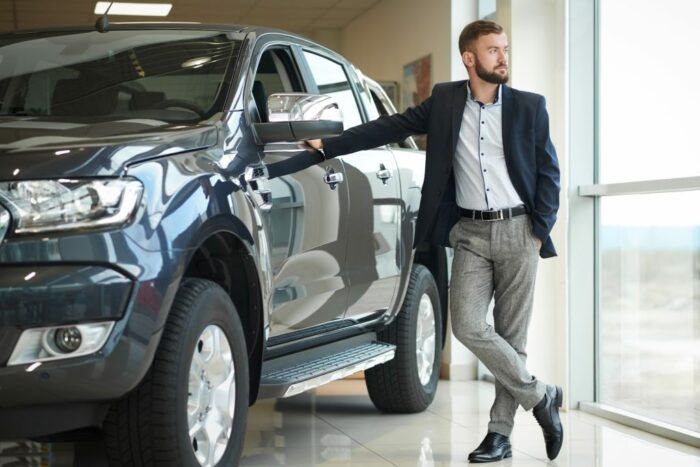 Ein junger Mann im Jacket posiert in einem Autohaus vor einem großen Auto (SUV). Die Fensterscheiben spiegeln sich auf dem Lack des Autos.