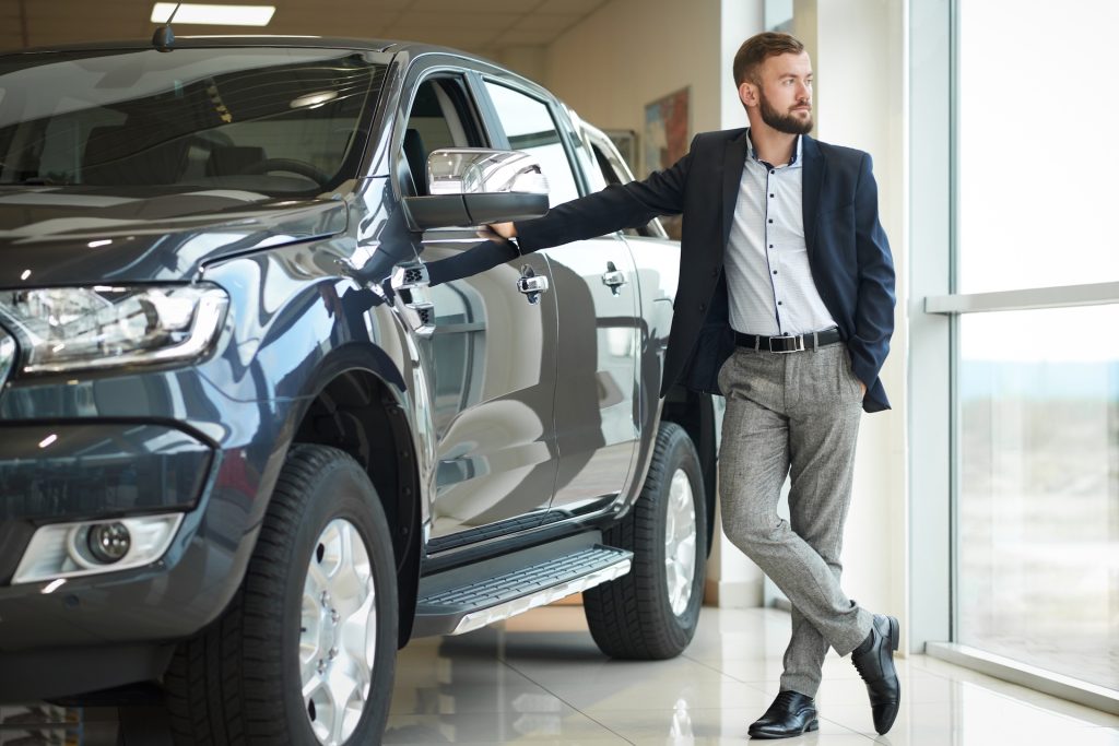 Ein junger Mann im Jacket posiert in einem Autohaus vor einem großen Auto (SUV). Die Fensterscheiben spiegeln sich auf dem Lack des Autos.