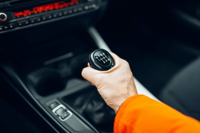 Ein Mensch sitzt im Auto und greift mit der rechten Hand den Ganghebel eines Autos mit Kupplung.