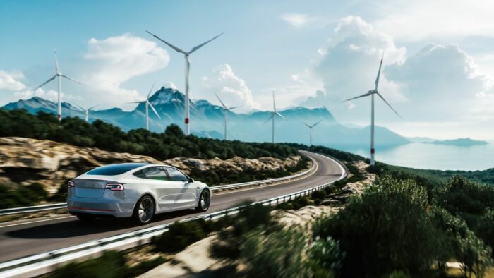 Ein E-Autos fährt durch eine karge Landschaft mit wenig Sträuchern. Im Hintergrund stehen Windräder vor einer Bergkulisse.