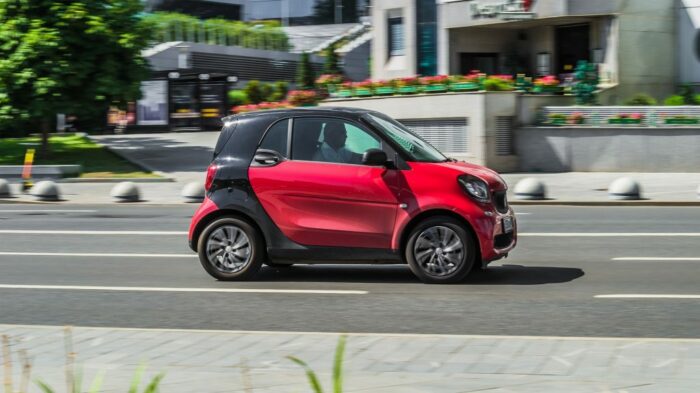 EIn roter Smart mit schwarzer B-Säule fährt auf einer Strasse an einem Haus mit Blumenkästen vorbei.