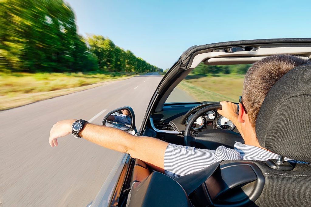 Ein Mann fährt mit einem Cabrio entspannt über eine leere Landstrasse und lässt seinen linken Arm über die Tür hinaus hängen.